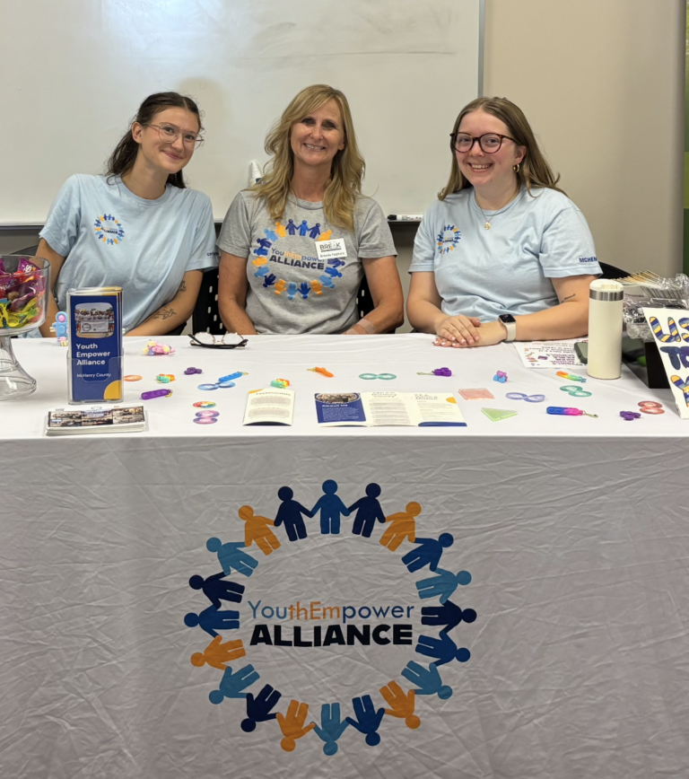 Three women sit at a table with the Youth Empower Alliance logo printed on the tablecloth.